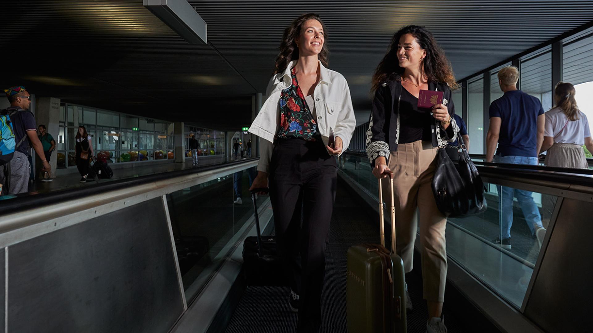 Twee vriendinnen gaan samen op reis. Ze lopen op Schiphol met hun paspoort in hun hand.