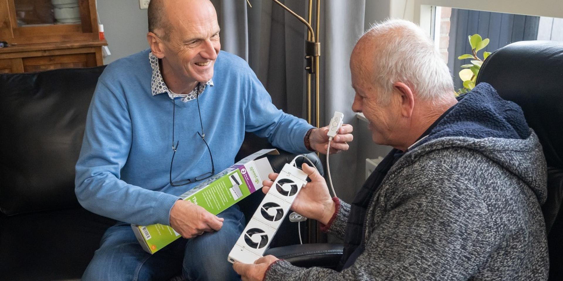 Energiecoach Aarnoud van der Deijl laat een radiatorventilator zien aan een oudere man bij wie hij op huisbezoek is.