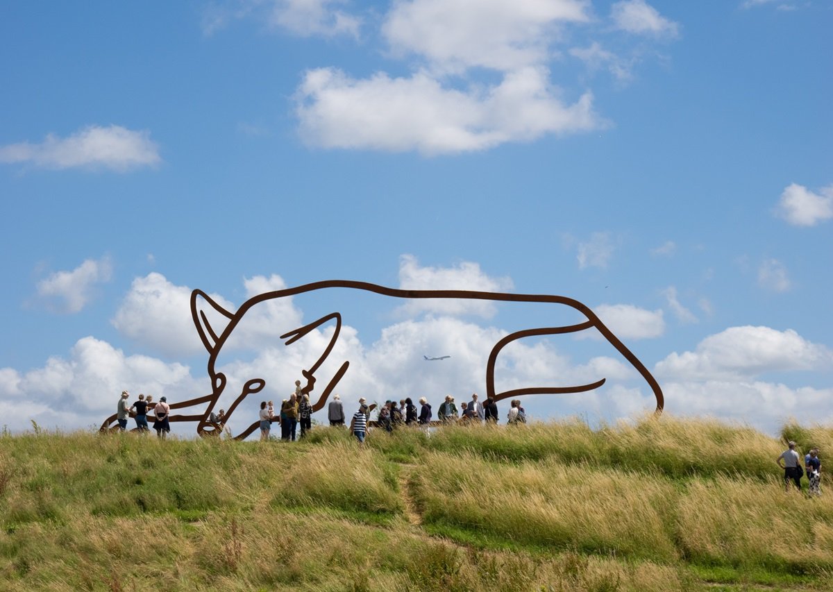 Silhouet van staal van een liggende wolf met een groepje mensen ervoor op een groen talud tegen een blauwe lucht met wolken.