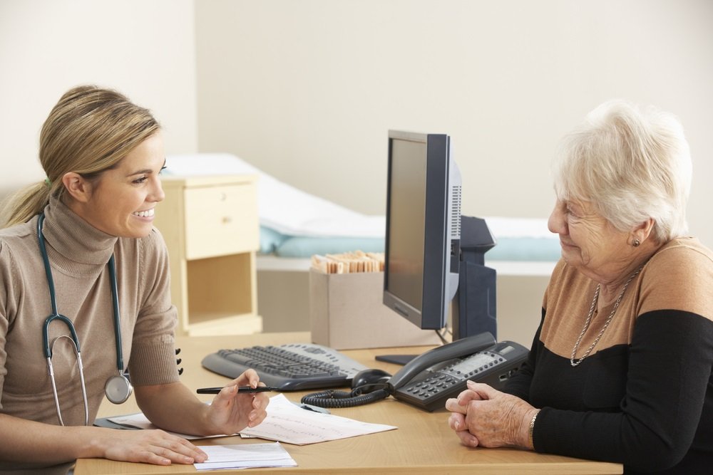 Een vrouw met een stethoscoop om haar nek achter een bureau in gesprek met een oudere dameeen oudere dame 